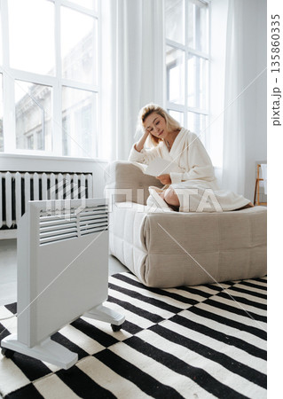 Young woman reading book near electric heater at home 135860335