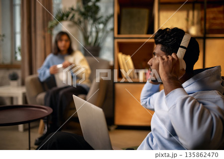 Young man wearing headphones using laptop in modern coworking environment with woman relaxing in background. Young man wearing headphones using laptop in modern coworking environment with woman relaxing in background. 135862470