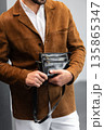 Stylish man in a brown suede blazer and white trousers leaning against a wall holding a small dark leather crossbody bag 135865347