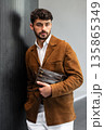 Stylish man in a brown suede blazer and white trousers leaning against a wall holding a small dark leather crossbody bag 135865349