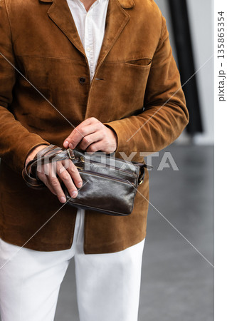 Stylish man in a brown suede blazer and white trousers leaning against a wall holding a small dark leather crossbody bag 135865354