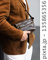 Stylish man in a brown suede blazer and white trousers leaning against a wall holding a small dark leather crossbody bag 135865356