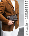 Stylish man in a brown suede blazer and white trousers leaning against a wall holding a small dark leather crossbody bag 135865357