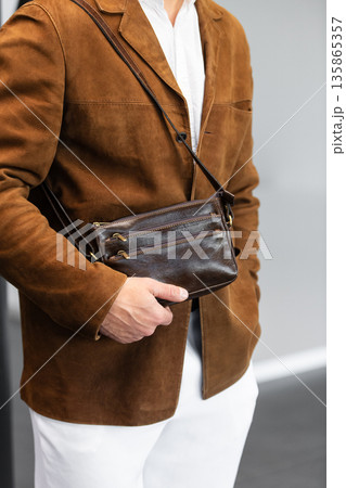 Stylish man in a brown suede blazer and white trousers leaning against a wall holding a small dark leather crossbody bag Stylish man in a brown suede blazer and white trousers leaning against a wall holding a small dark leather crossbody bag 135865357