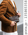 Stylish man in a brown suede blazer and white trousers leaning against a wall holding a small dark leather crossbody bag 135865358