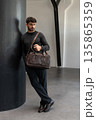 Confident man in sunglasses and charcoal knitwear holding a dark brown leather briefcase while leaning against a textured pillar in a modern minimalist interior. 135865359