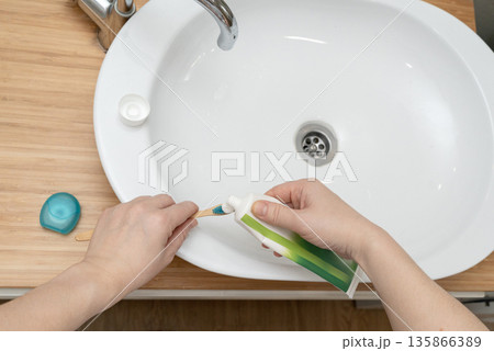 Top view of hands applying toothpaste on toothbrush over white bathroom sink. Daily dental hygiene routine, clean minimalist bathroom and healthy lifestyle concept Top view of hands applying toothpaste on toothbrush over white bathroom sink. Daily dental hygiene routine, clean minimalist bathroom and healthy lifestyle concept 135866389
