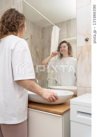 Woman brushing teeth with a bamboo toothbrush, smiling at her reflection in the mirror. Daily dental hygiene routine Woman brushing teeth with a bamboo toothbrush, smiling at her reflection in the mirror. Daily dental hygiene routine 135866390