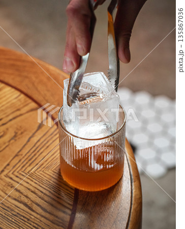 Bartender placing ice cube into amber cocktail Bartender placing ice cube into amber cocktail 135867036
