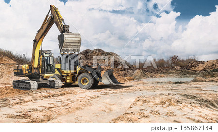 A loader and crawler excavator work at a construction site, moving soil for future work. The partially cloudy sky creates a contrast with the landscape in the background 135867134