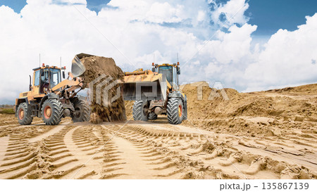 Two wheel loaders. Heavy equipment is working on sandy soil at a construction site. Track marks are visible in the sand, and a large pile of sand is visible in the background Two wheel loaders. Heavy equipment is working on sandy soil at a construction site. Track marks are visible in the sand, and a large pile of sand is visible in the background 135867139