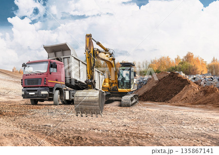 Loader moves earth while excavator digs at construction site. A truck is present, unloading materials near rental equipment under a clear sky and clouds 135867141