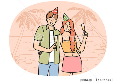 Honeymoon happy couple drinking champagne, standing on beach of sunny resort, wearing birthday caps 135867331