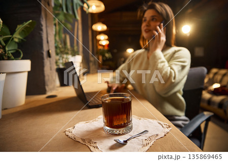 Glass of black coffee on wooden table with woman talking on smartphone and laptop 135869465