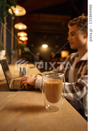 Double wall latte glass on table with man working on laptop. Double wall latte glass on table with man working on laptop. 135869466