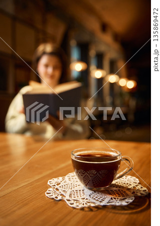 Glass coffee cup on wooden table with blurred reading woman. Glass coffee cup on wooden table with blurred reading woman. 135869472