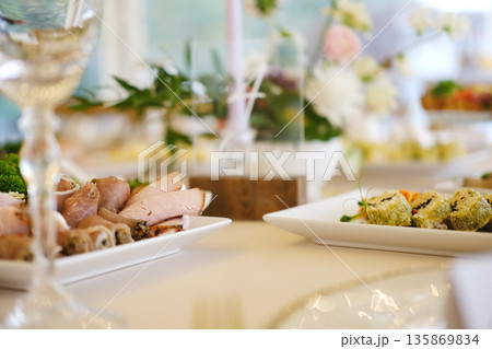 Platter of sliced meats and sushi rolls on white plates with greenery, crystal glassware, and floral decorations on dining table during event setup 135869834