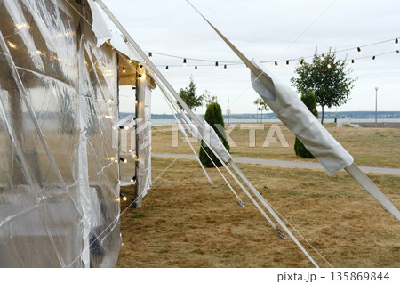 Tent with clear plastic walls and string lights set up on grass field with trees and pathway in background under cloudy sky during evening Tent with clear plastic walls and string lights set up on grass field with trees and pathway in background under cloudy sky during evening 135869844