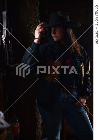 Cowgirl in a wide brim hat leans against a dark stable, exuding confidence at the ranch 135870055