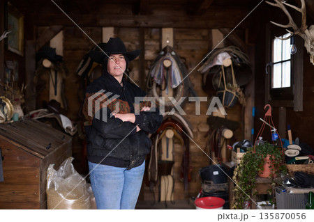 Cowgirl in a hat stands with arms crossed inside a rustic tack room filled with saddles Cowgirl in a hat stands with arms crossed inside a rustic tack room filled with saddles 135870056