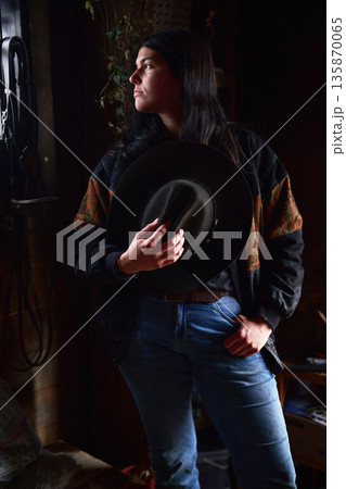 Portrait of a woman in a casual outfit holding a hat in moody indoor lighting Portrait of a woman in a casual outfit holding a hat in moody indoor lighting 135870065