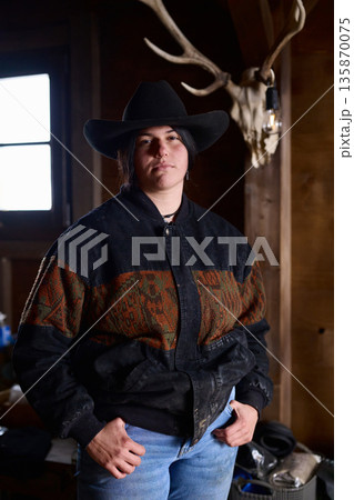 Cowgirl in a wide brimmed hat stands confidently in a rustic ranch interior Cowgirl in a wide brimmed hat stands confidently in a rustic ranch interior 135870075