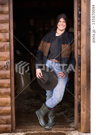 Smiling cowgirl stands in rustic barn doorway wearing jacket, jeans, and hat 135870094