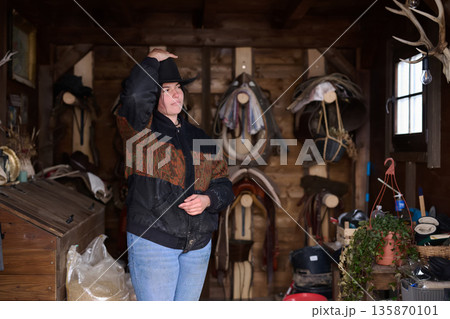 Cowgirl in a rustic barn adjusts her hat among saddles and tack, ready for work 135870101