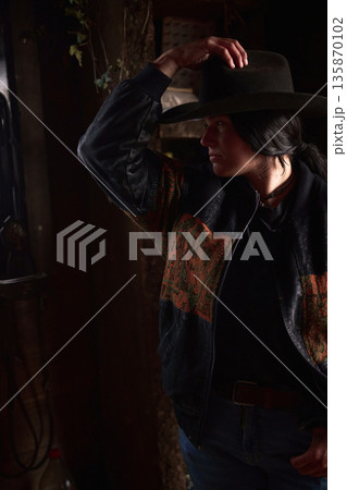 Cowgirl in a dark ranch interior adjusts her hat, wearing a patterned jacket and jeans 135870102