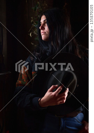 Portrait of a woman with dark hair holding a hat in a moody intimate studio setting 135870105