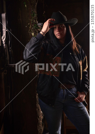 Cowgirl in a rustic ranch setting adjusting her hat in denim jacket and jeans Cowgirl in a rustic ranch setting adjusting her hat in denim jacket and jeans 135870135