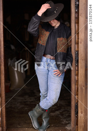 Cowgirl in denim, hat and boots standing in a rustic doorway by a wooden barn 135870144
