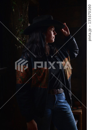 Cowgirl in a wide brim hat and patterned jacket stands by a doorway in moody, rustic light Cowgirl in a wide brim hat and patterned jacket stands by a doorway in moody, rustic light 135870160