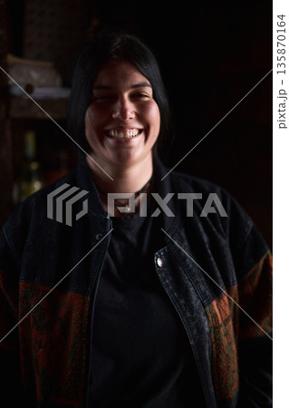 Smiling woman in a dark jacket stands against a dimly lit background, conveying warmth and casual style 135870164