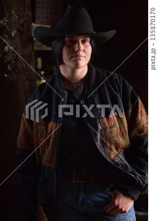 Cowgirl in a dark, stylish jacket and wide-brim hat, standing at a rustic ranch setting Cowgirl in a dark, stylish jacket and wide-brim hat, standing at a rustic ranch setting 135870170