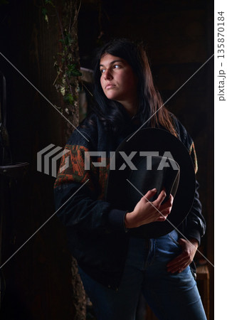 A thoughtful woman in a dark, rustic setting holds a wide-brimmed hat A thoughtful woman in a dark, rustic setting holds a wide-brimmed hat 135870184