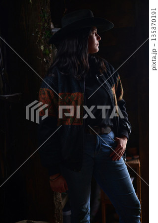 Cowgirl in wide hat and denim, standing in a moody ranch setting with a confident pose Cowgirl in wide hat and denim, standing in a moody ranch setting with a confident pose 135870191