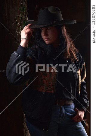 Cowgirl in Wide Hat and Dark Jacket Stands by Rustic Wood Wall, Moody Western Portrait 135870205