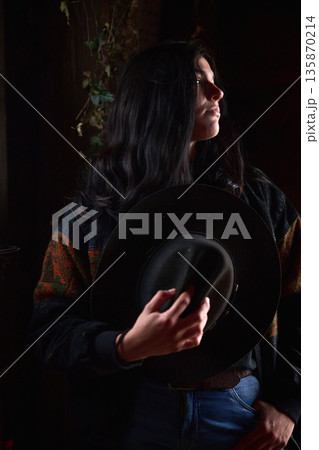 Moody portrait of a woman holding a wide brim hat in a dimly lit setting Moody portrait of a woman holding a wide brim hat in a dimly lit setting 135870214