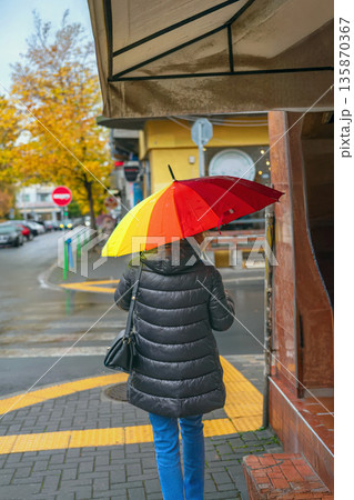 Woman in black coat walking on rainy autumn city street holding bright rainbow umbrella. Concept of urban solitude, autumn rain, urban fall mood, seasonal weather Woman in black coat walking on rainy autumn city street holding bright rainbow umbrella. Concept of urban solitude, autumn rain, urban fall mood, seasonal weather 135870367