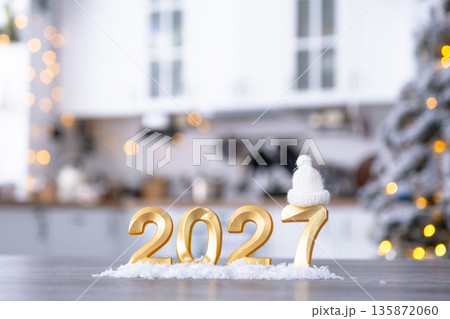 Golden number 2027 on table in background of cozy white kitchen with Christmas tree and Christmas decor with garland lights. Housing, interior, renovation, mortgage, rent in New Year 135872060