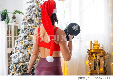 Woman in sports uniform and Santa hat is engaged in fitness and strength training with dumbbells at home in Christmas interior with Christmas tree. Recovery of athletic form after New Year Woman in sports uniform and Santa hat is engaged in fitness and strength training with dumbbells at home in Christmas interior with Christmas tree. Recovery of athletic form after New Year 135872711