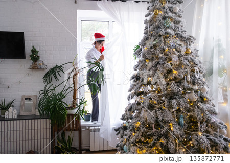A woman hang up white curtain on the window, preparation for the Christmas and New Year, fairy lights in the interior of a house with a Christmas tree 135872771