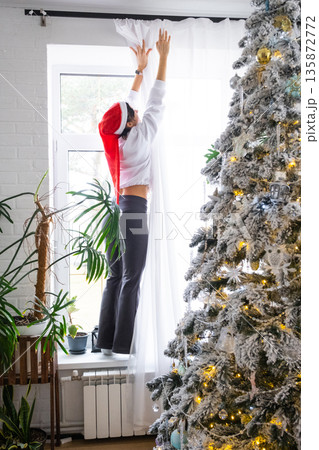 A woman hang up white curtain on the window, preparation for the Christmas and New Year, fairy lights in the interior of a house with a Christmas tree 135872772