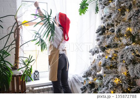 Woman in a Santa hat and apron cleans window near Christmas tree, preparing for the New Year, decorating and tidying up the house Woman in a Santa hat and apron cleans window near Christmas tree, preparing for the New Year, decorating and tidying up the house 135872788