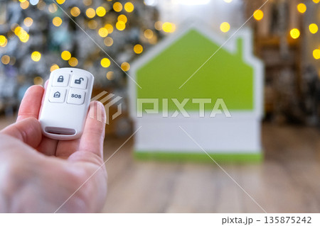 Security System Keychain in hand on background fairy lights Christmas tree and tiny house gift for New Year, Christmas. Home security, alarm system, SOS signal, protection from thieves Security System Keychain in hand on background fairy lights Christmas tree and tiny house gift for New Year, Christmas. Home security, alarm system, SOS signal, protection from thieves 135875242