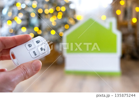 Security System Keychain in hand on background fairy lights Christmas tree and tiny house gift for New Year, Christmas. Home security, alarm system, SOS signal, protection from thieves 135875244
