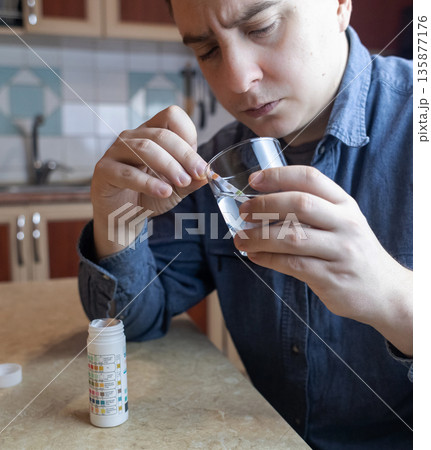 A man in the kitchen tests tap water quality with a test strip. Home testing for drinking water safety, pH, chlorine, and chemical contaminants. 135877176