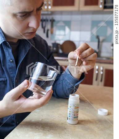 A man in the kitchen tests tap water quality with a test strip. Home testing for drinking water safety, pH, chlorine, and chemical contaminants. 135877178