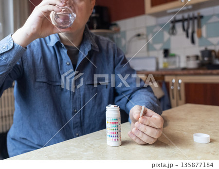 A man in the kitchen tests tap water quality with a test strip. Home testing for drinking water safety, pH, chlorine, and chemical contaminants. 135877184